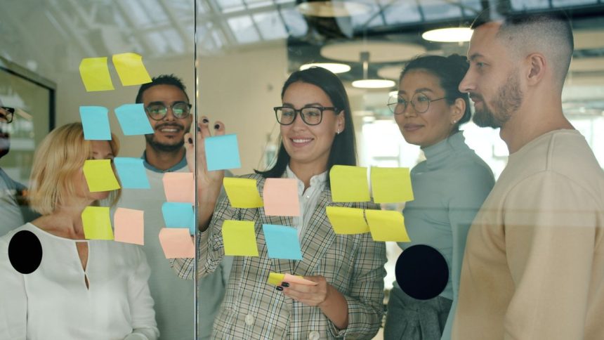 Profissionais colaboram em um escritório, organizando ideias com notas adesivas em uma parede de vidro.