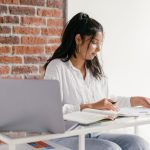 Estudante usa um laptop e consulta um caderno em uma mesa durante os estudos em casa.