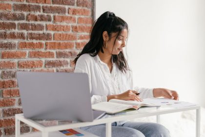 Estudante usa um laptop e consulta um caderno em uma mesa durante os estudos em casa.